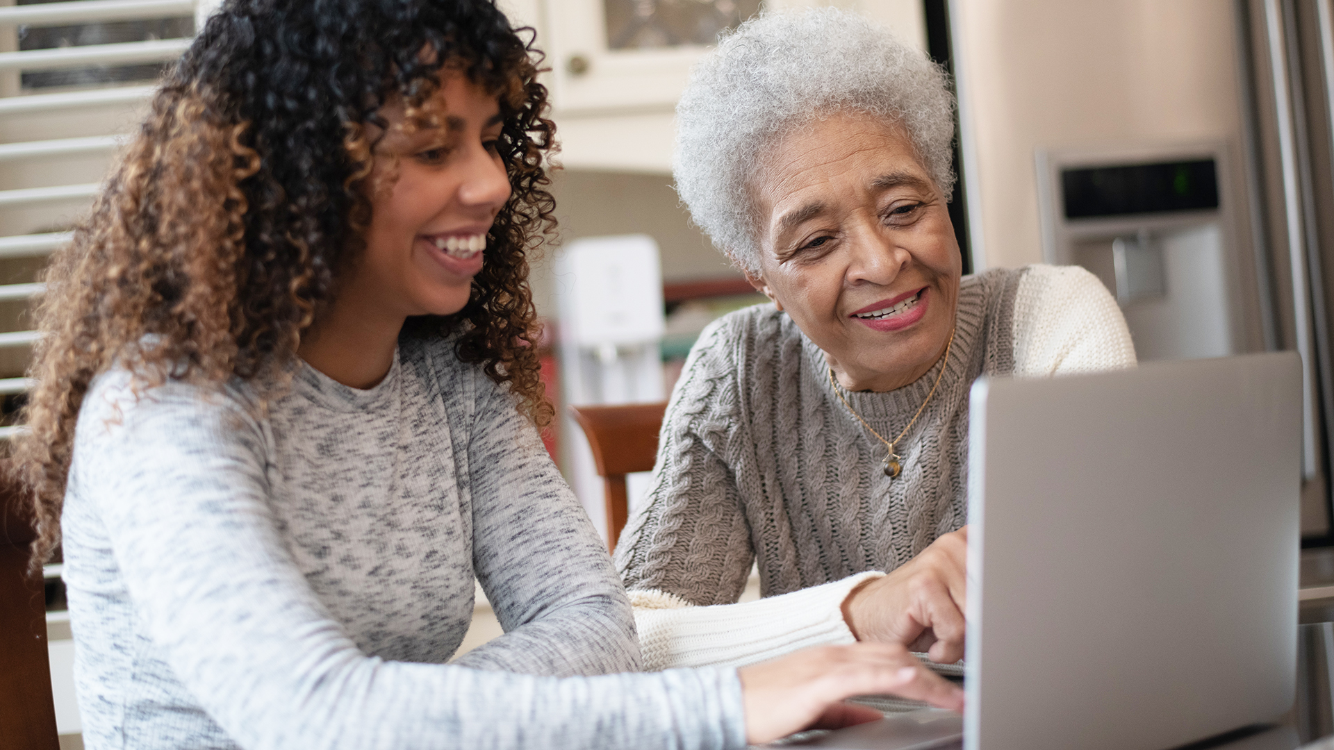 A young mature woman of African decent, shows her elderly mother how to video chat with family on a laptop.  They are both seated at the kitchen table, dressed casually and smiling as they enjoy the conversation with family far away.